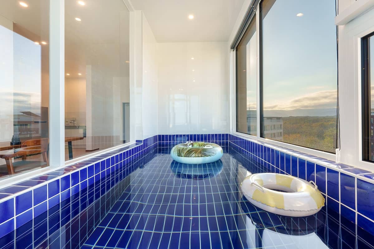 A modern bathroom with a blue-tiled jacuzzi, a yellow and white inflatable ring, and a view of the cityscape through large windows.