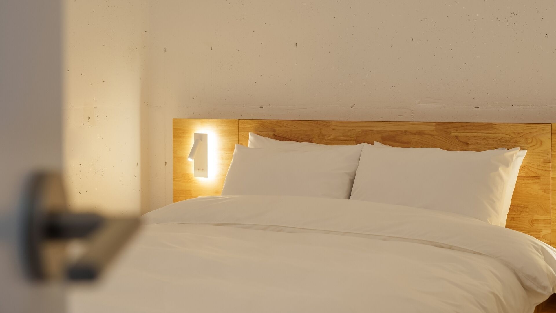 A neatly made bed with white linens, a wooden headboard, and a wall-mounted light fixture.