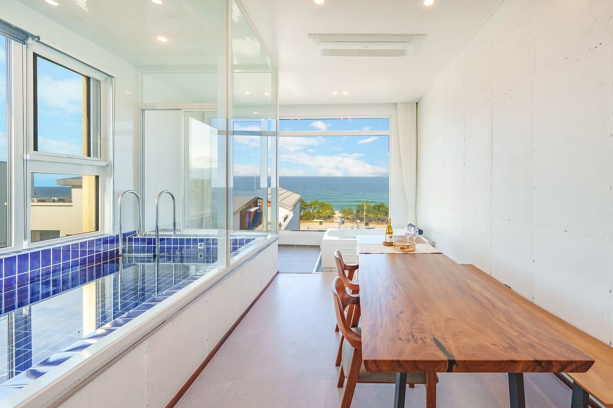A modern kitchen with a large window overlooking the sea, a wooden dining table, and a blue-tiled sink.
