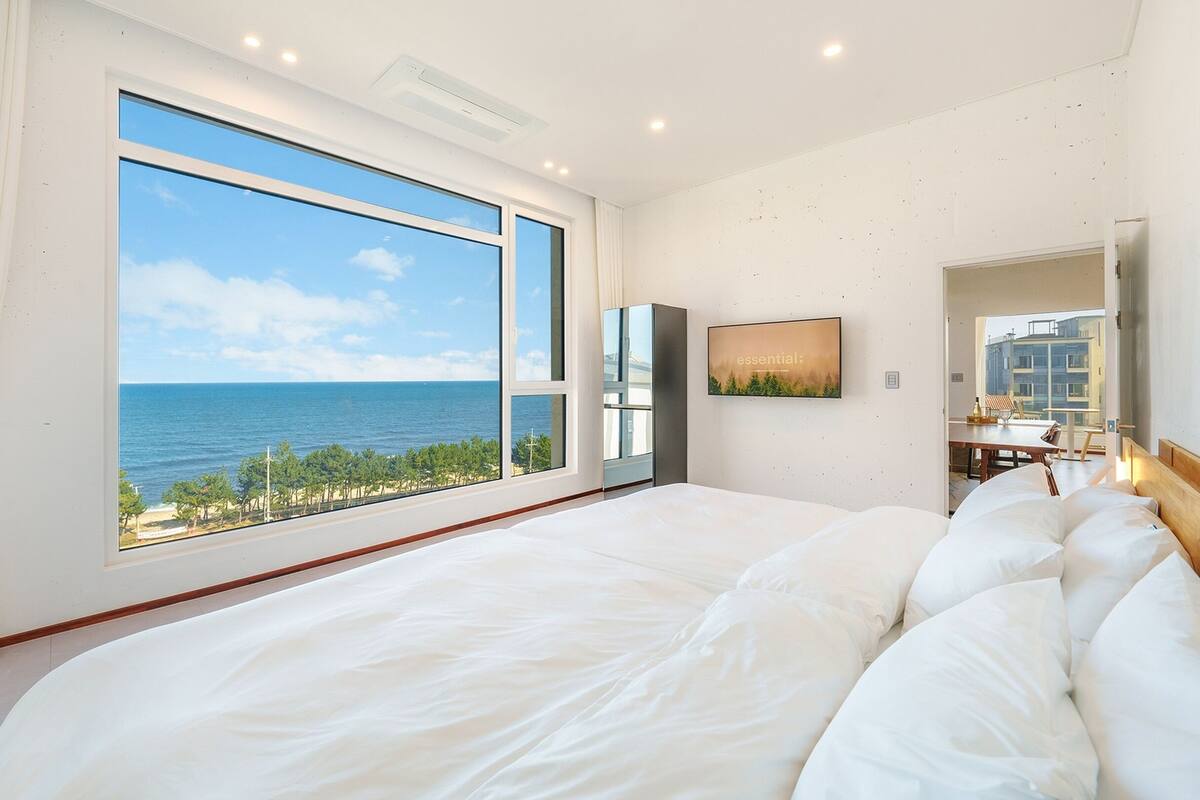 A large bed with white linens, a large window with a view of the ocean, and a flat-screen TV on the wall.