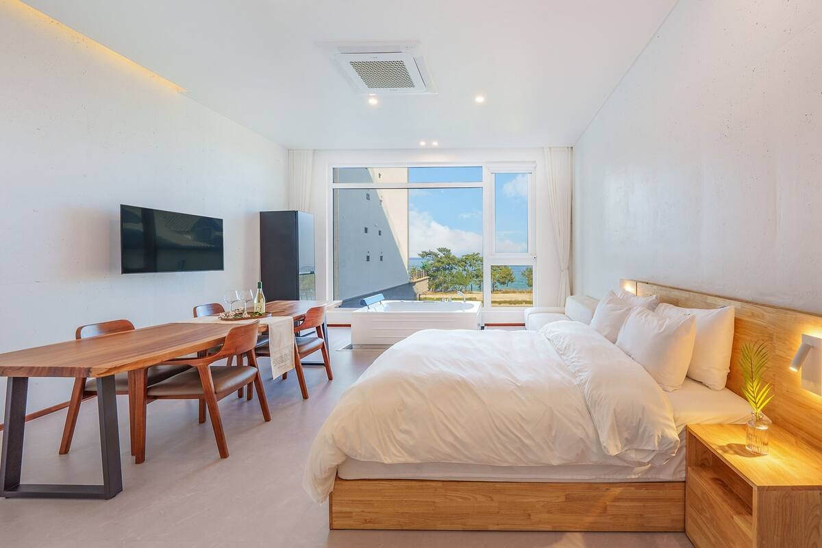 A modern bedroom with a large bed, a wooden nightstand, a desk with chairs, a flat-screen TV, and a large window offering a view of the outdoors.