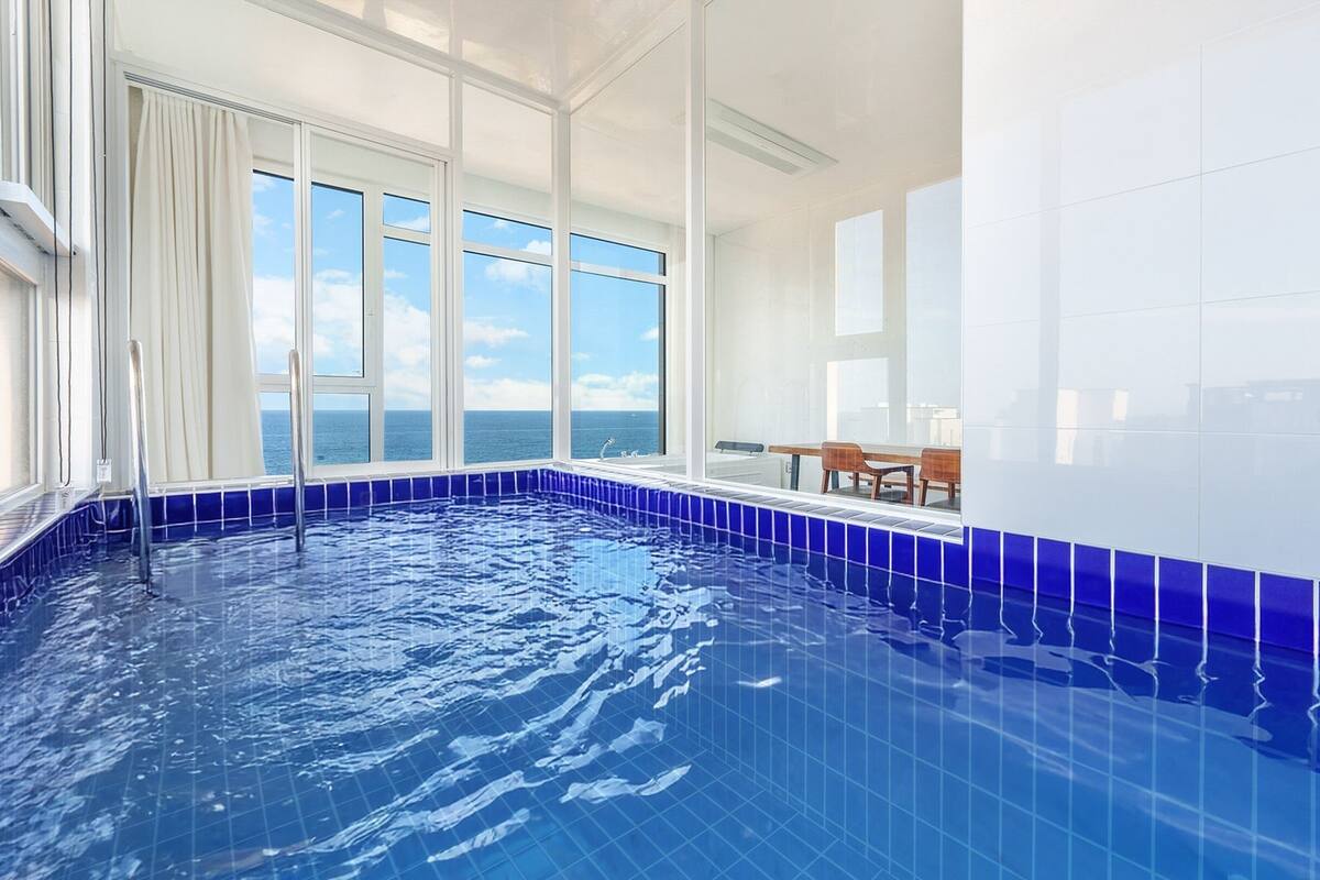 A large indoor swimming pool with clear blue tiles, a view of the ocean through large windows, and a seating area with a table and chairs.