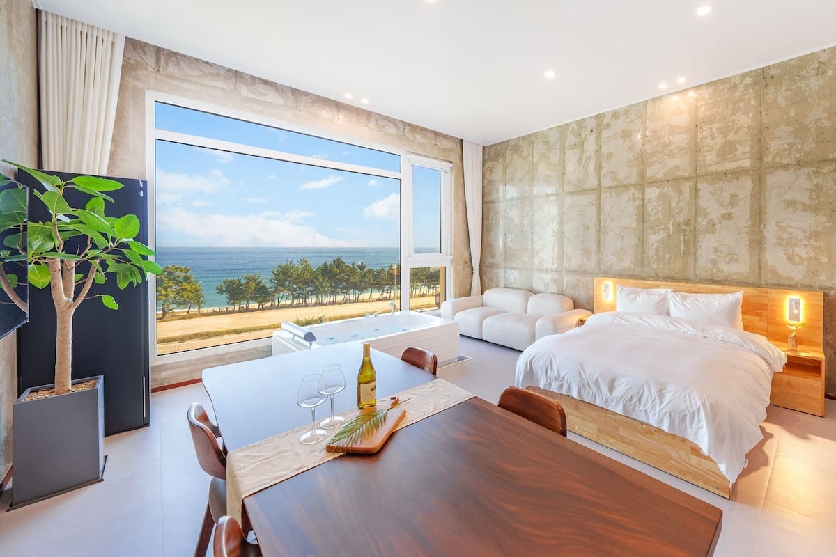 A modern hotel room with a large window offering a sea view, a cozy bed, a dining area, and a built-in refrigerator.