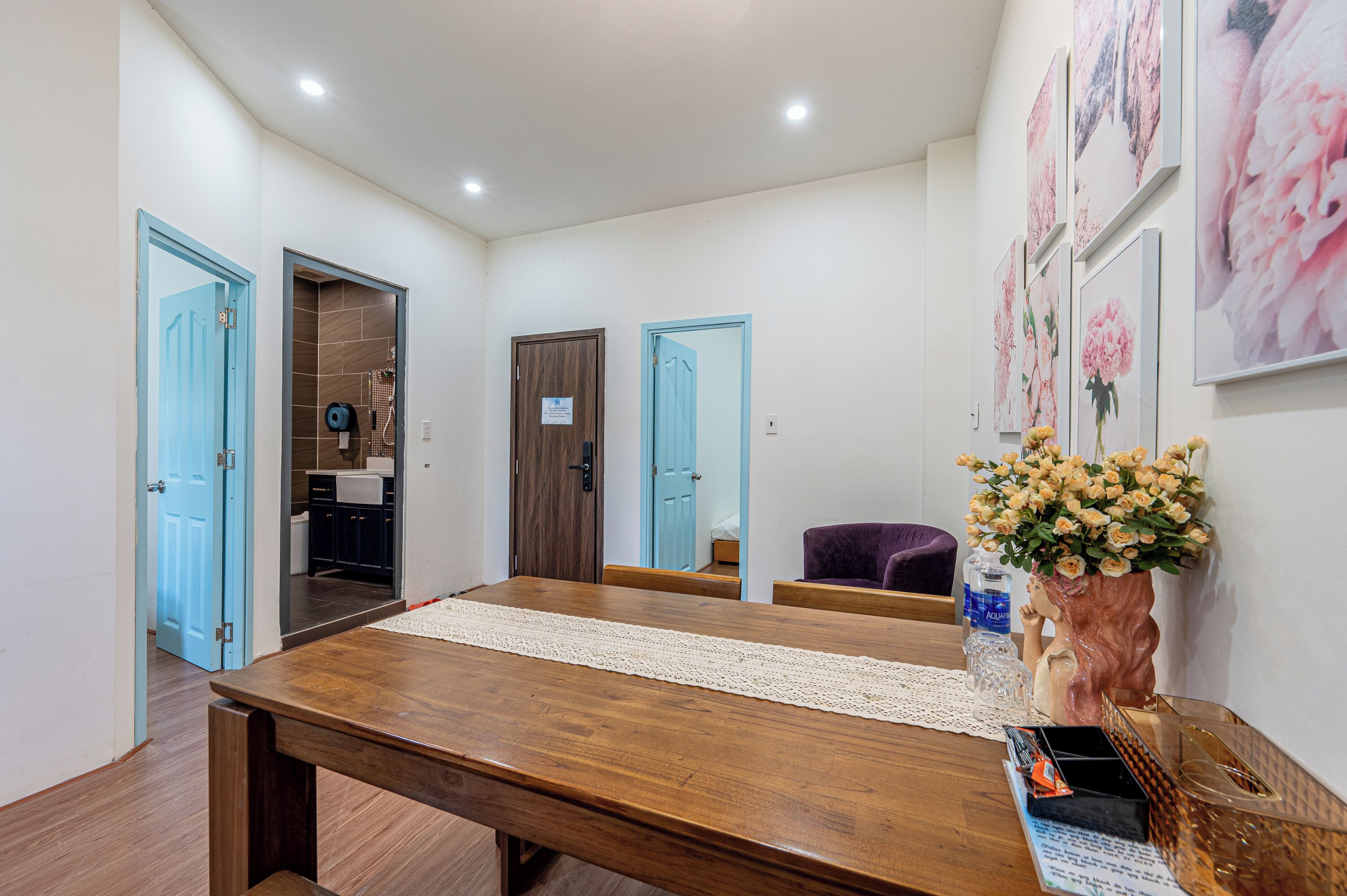 A spacious room with a wooden dining table, a purple chair, and a vase of flowers. There are three doors leading to other rooms.