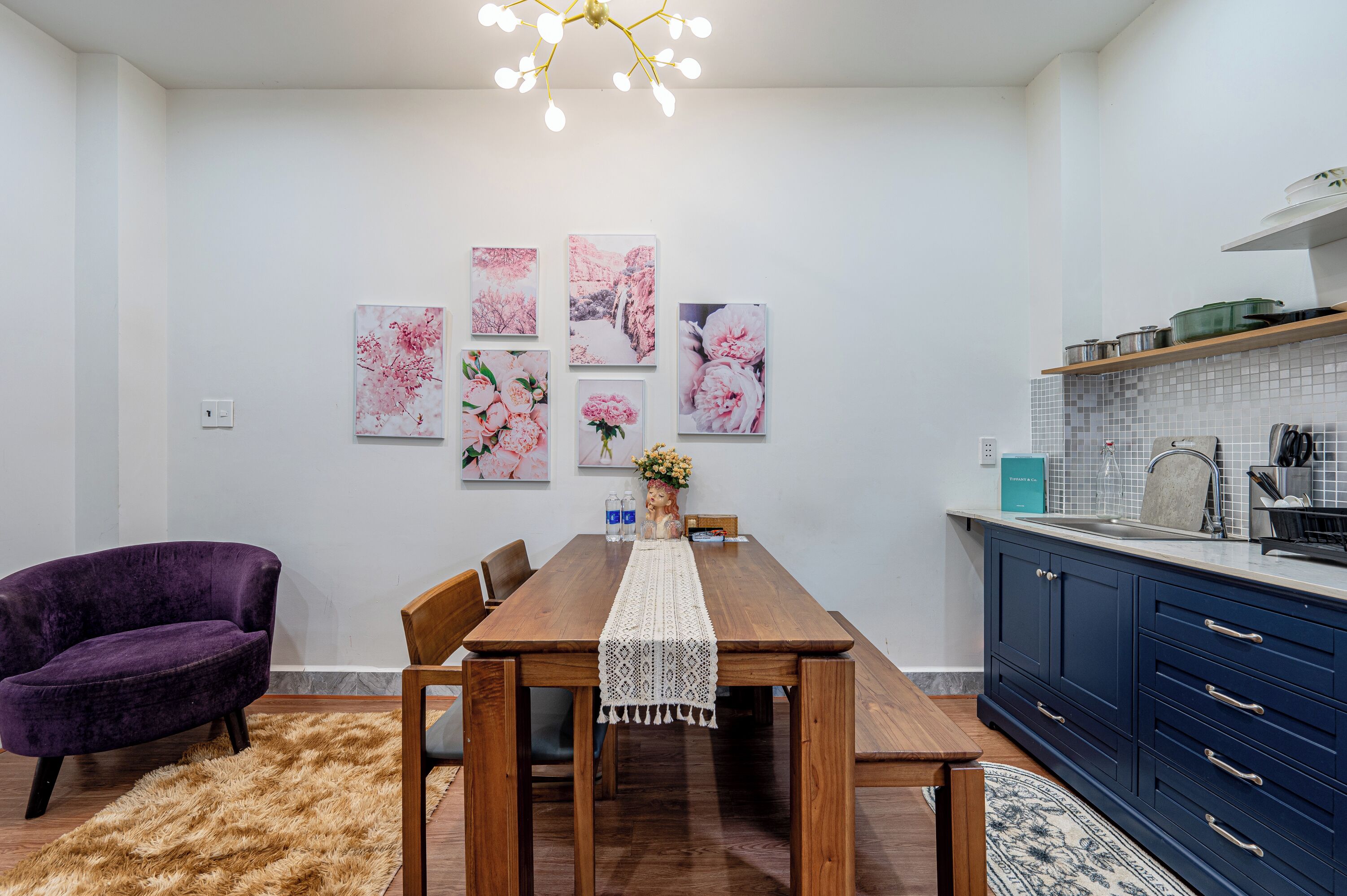 A dining area with a wooden table, purple chair, and a rug. There are wall-mounted pictures and a kitchen area with cabinets and a countertop.
