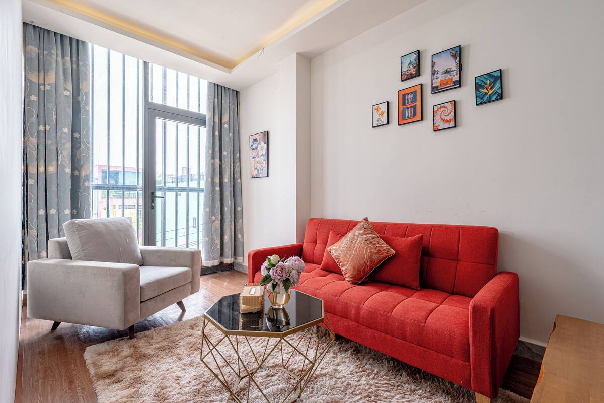 A modern living room with a red sofa, a white armchair, and a glass coffee table. There are framed artworks on the wall and a window with curtains.