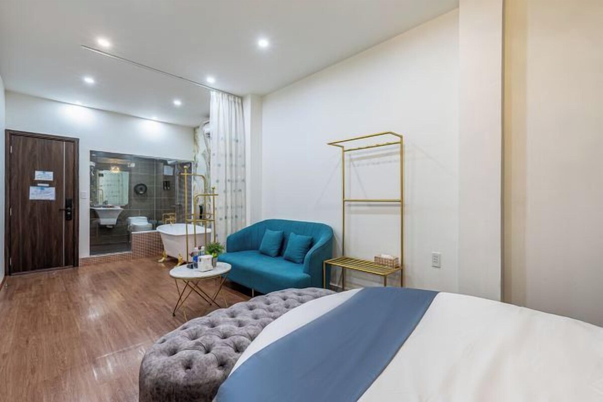 A modern hotel room with a bed, a blue sofa, a tufted ottoman, a small round table with a plant, and a gold-framed shelf.