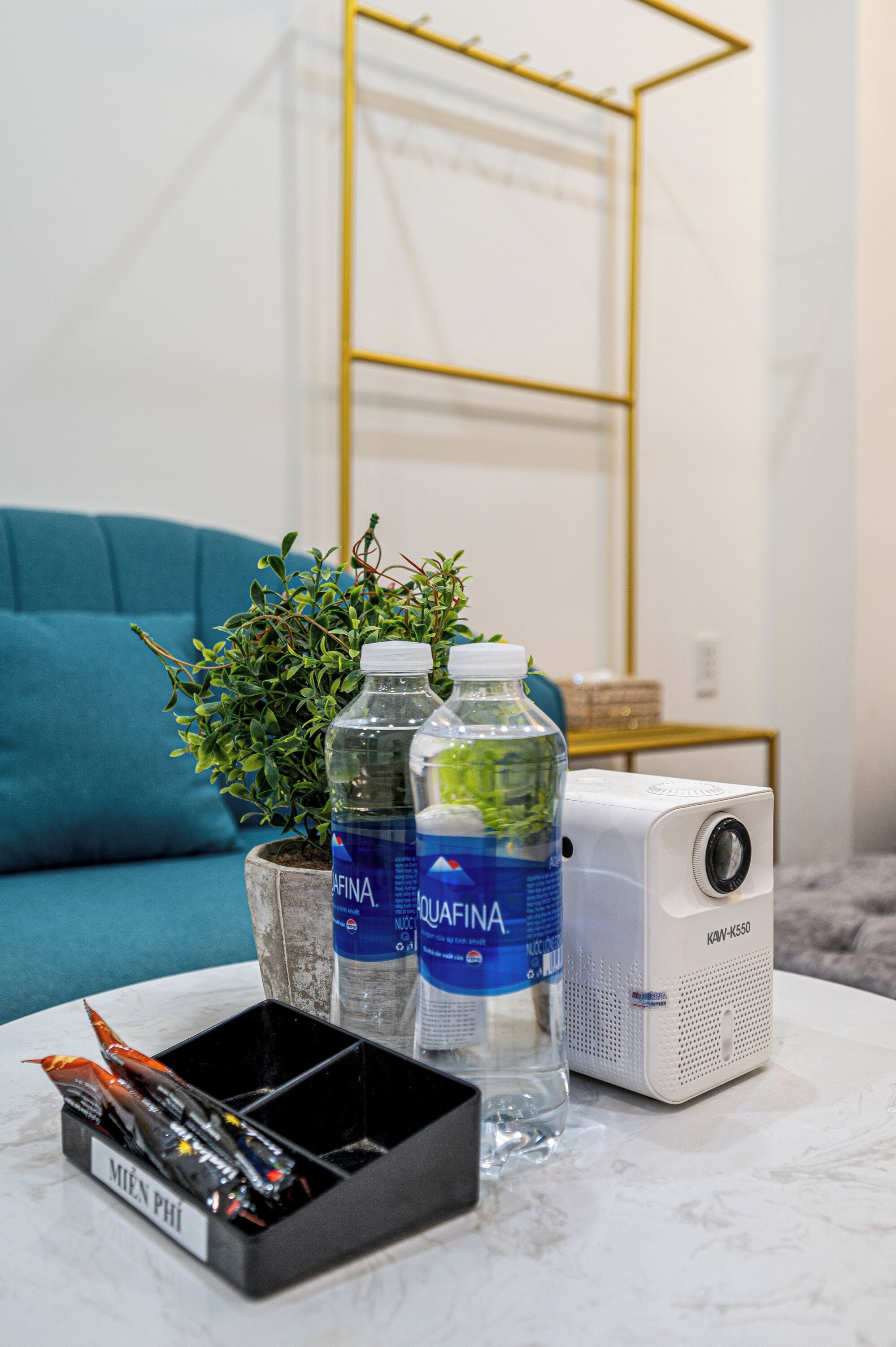 Een kamer met een blauwe bank, een witte salontafel, twee waterflessen, een kleine potplant en een witte speaker.