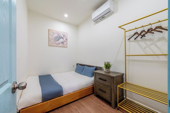 Un dormitorio con cama, mesita de noche, aire acondicionado de pared, un cuadro enmarcado y perchero.
