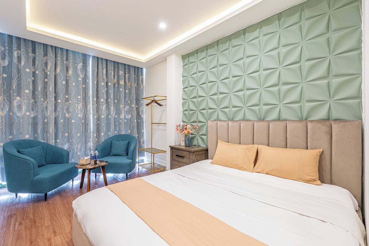 A modern hotel room with a large bed, two armchairs, a side table with a vase of flowers, and a wardrobe.