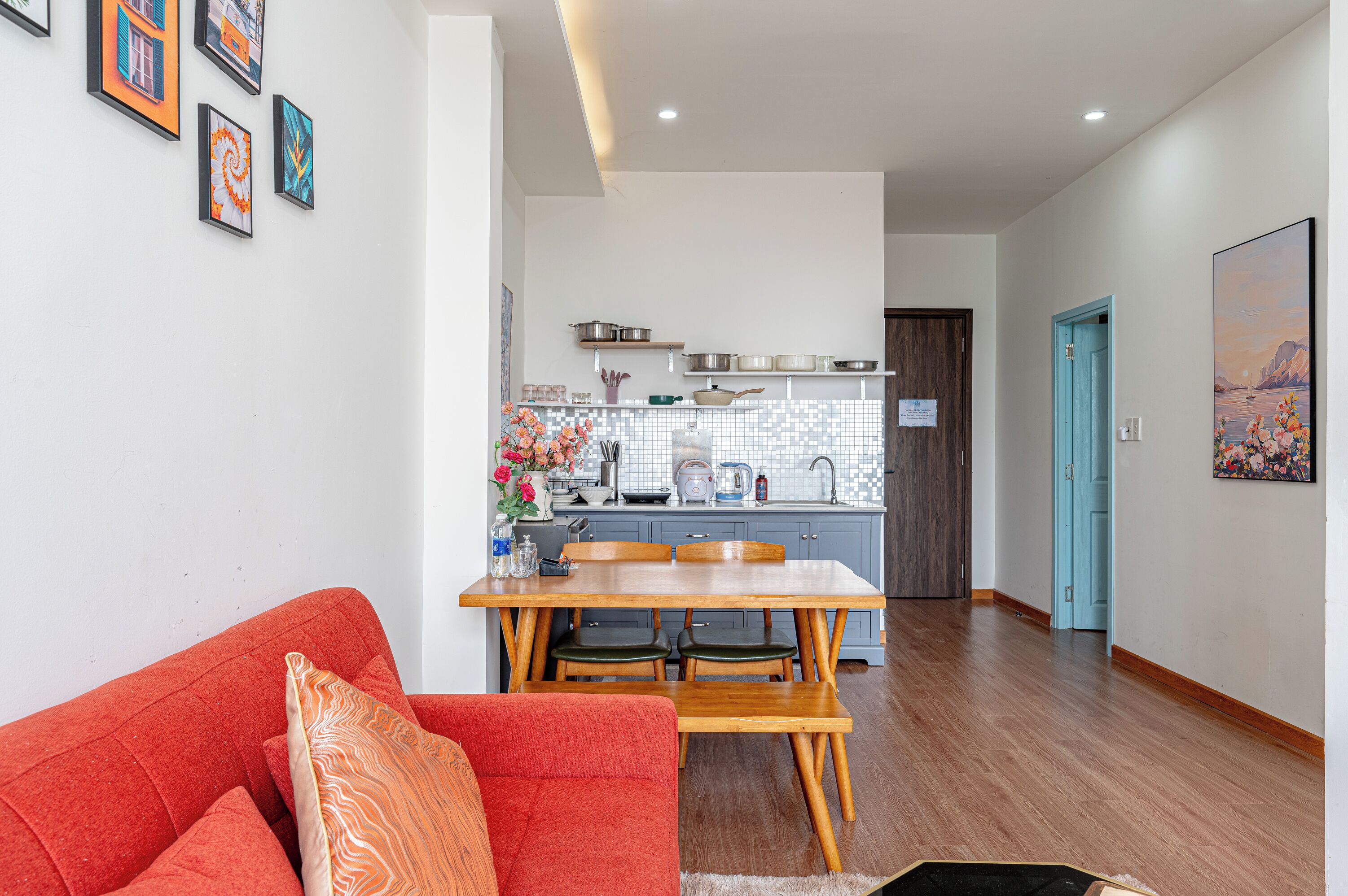A modern kitchen with wooden cabinets, a dining table with chairs, and a red sofa with patterned cushions.