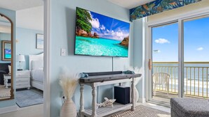 A room with a television mounted on the wall, a wooden table with a speaker, a white chair, and a view of the beach through a sliding glass door. - Bay Watch 509 (North Myrtle Beach)