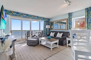 A living room with a sofa, coffee table, and a television. There is a balcony with a view of the ocean.