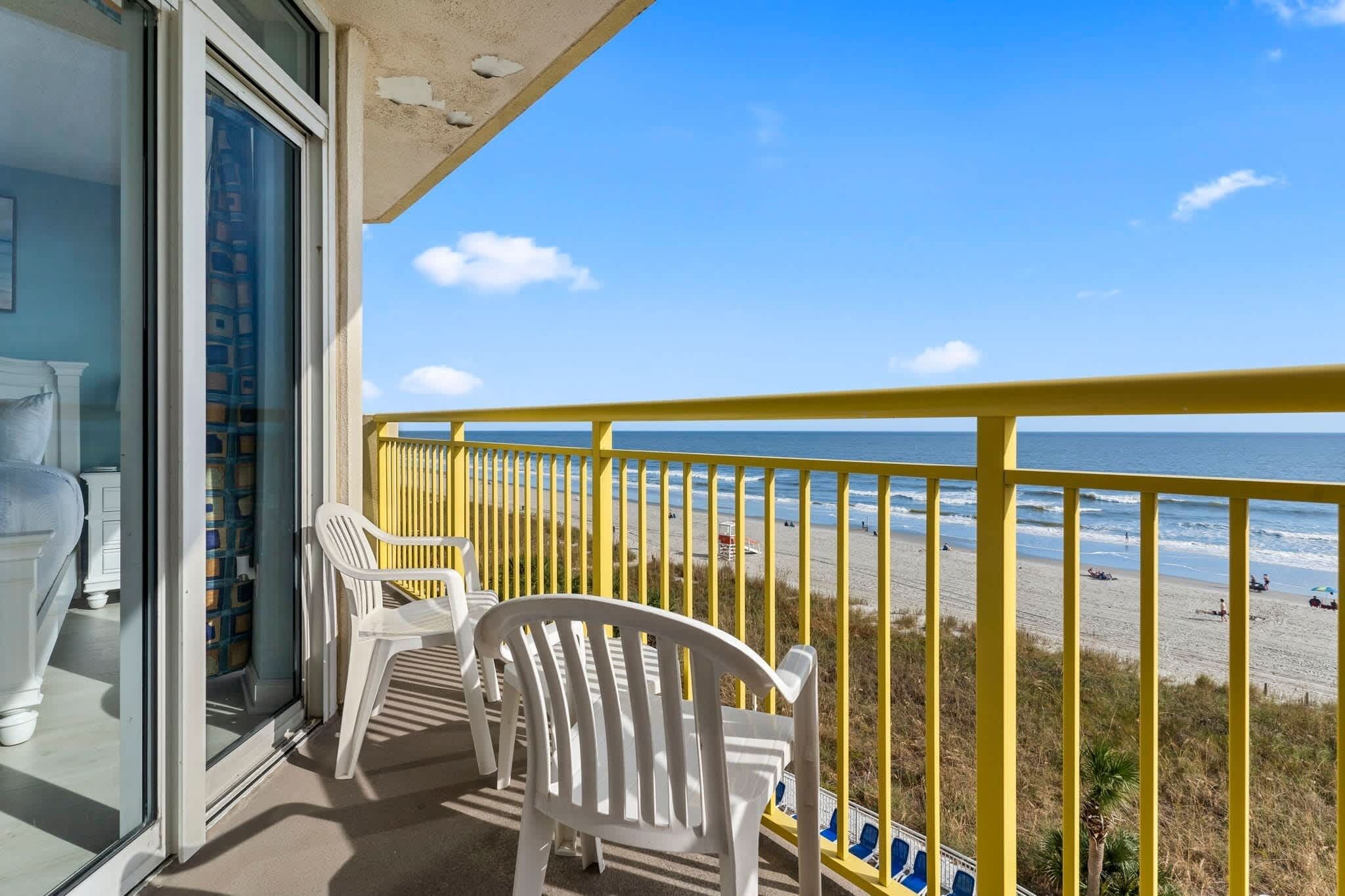 Ein Balkon mit gelbgerahmter Brüstung, weißen Plastikstühlen und Blick auf den Strand.