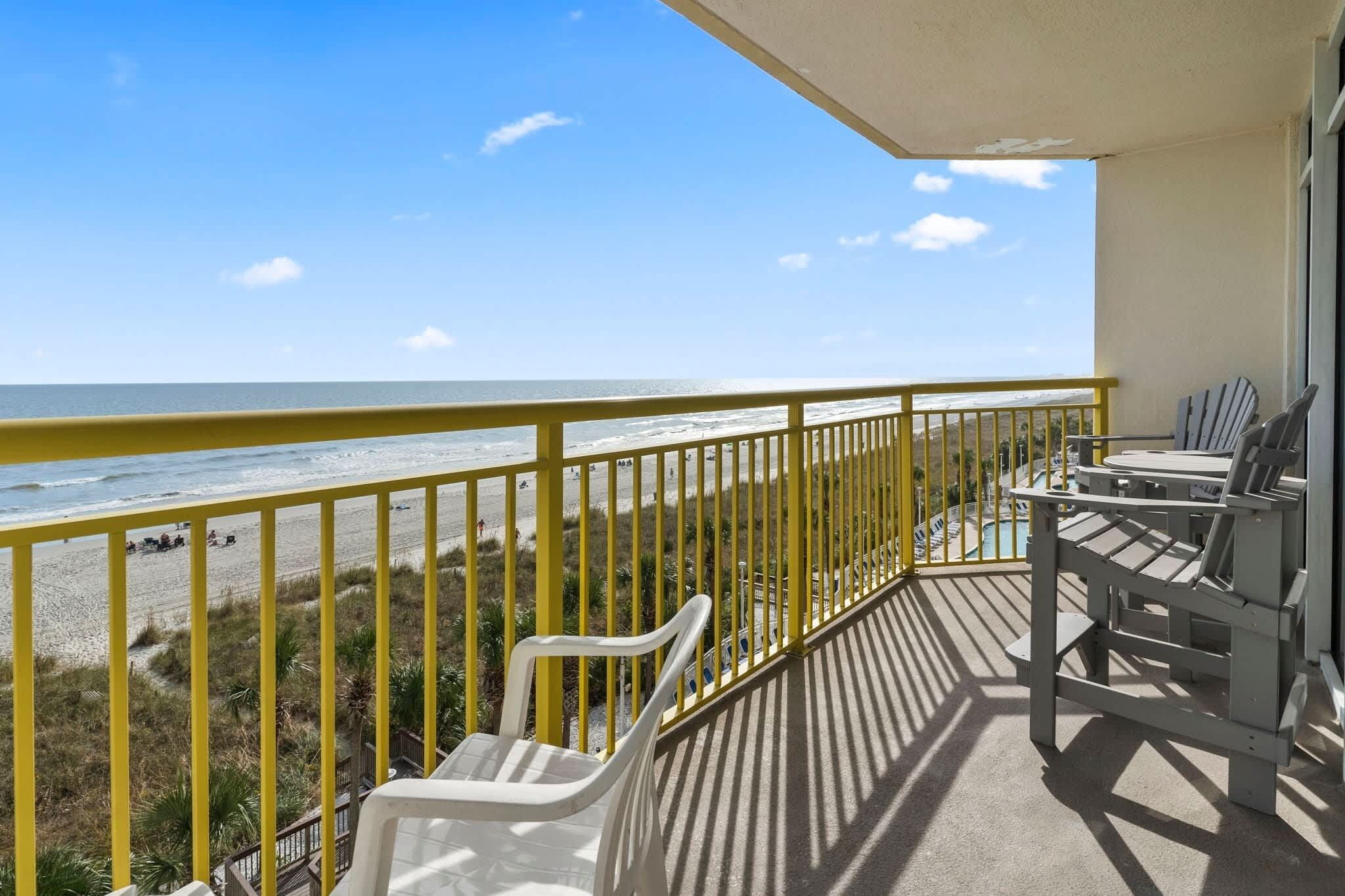 Ein Balkon mit Blick auf Strand und Meer, ein gelander und ein Tisch mit Stühlen.