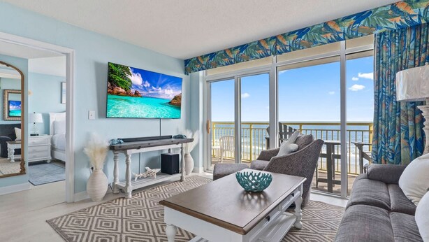 A living room with a large TV, a sofa, a coffee table, and a view of the ocean through large windows. - Bay Watch 509 (North Myrtle Beach)