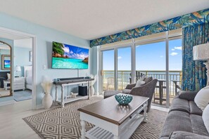 A living room with a large TV, a sofa, a coffee table, and a view of the ocean through large windows.
