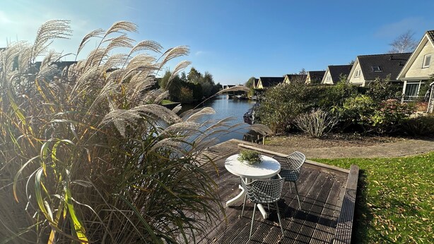 A riverside view with a dining area featuring a table and chairs, surrounded by lush greenery and a row of houses.