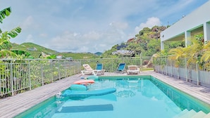 A rectangular swimming pool with a wooden deck, surrounded by a metal fence. There are lounge chairs and umbrellas along the poolside. - Seemingly unobstructed sea and marina view (Oyster Pond)