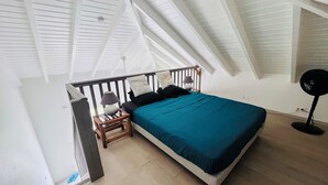 A bedroom with a sloped ceiling, a bed with a teal bedspread, a bedside table with a lamp, and a standing fan. - Seemingly unobstructed sea and marina view (Oyster Pond)