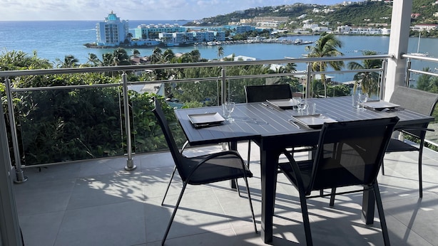 A balcony with a table and chairs overlooking a marina and coastal town. - Seemingly unobstructed sea and marina view (Oyster Pond)