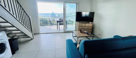 A living room with a sofa, a television, a coffee table, and a washing machine. There is a balcony with a view of the sea and buildings.