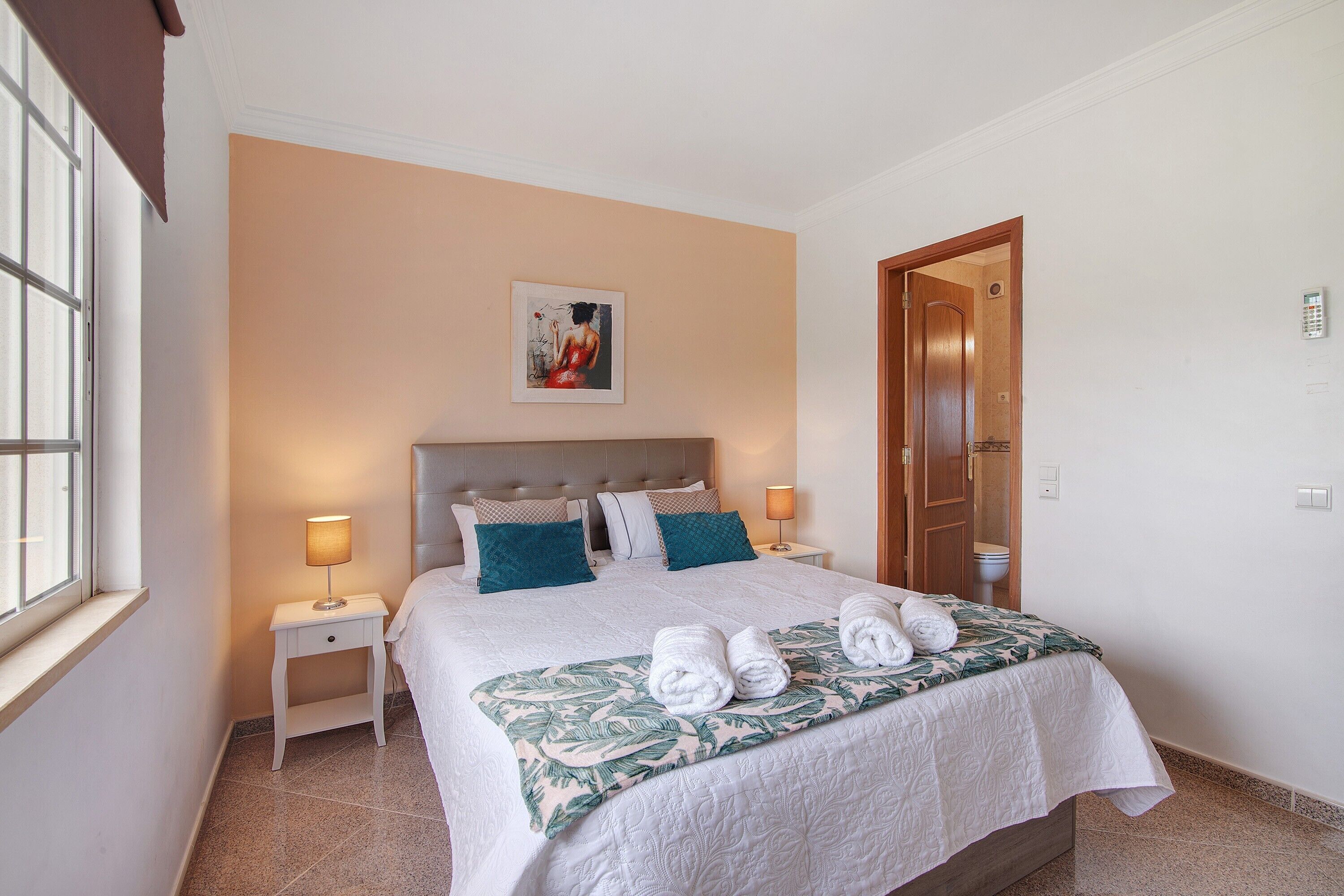 A neatly made bed with a floral bedspread, two bedside lamps, and a painting on the wall.