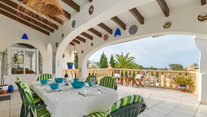 Een buiten eethoek met een tafel gedekt voor vier personen, omringd door gestreepte stoelen, onder een overdekt terras met decoratieve borden aan het plafond.
