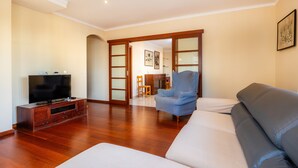 Una sala de estar con televisor, sofá y sillón, piso de madera, y una puerta que da a otra habitación.