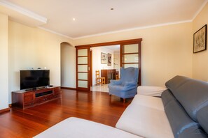 Un salon comprenant un téléviseur, un canapé et un fauteuil, un plancher en bois et une porte donnant sur une autre pièce.