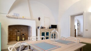 Een eetruimte met een open haard van baksteen, witte stoelen, een houten tafel en een televisie op een kast.