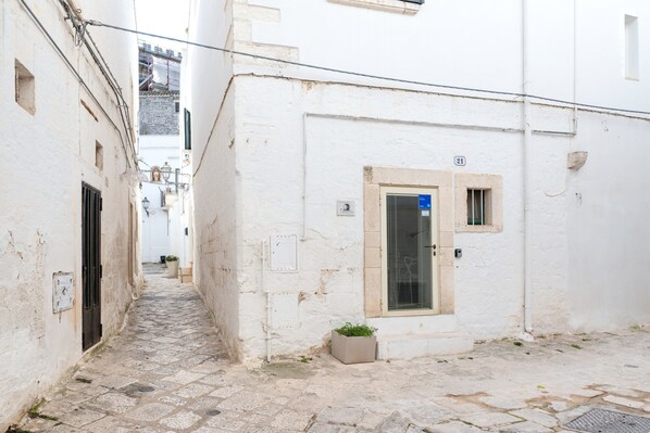 Une ruelle étroite pavée de pierres, bordée de bâtiments blancs, avec une porte dotée d’une fenêtre et un petit jardinière.