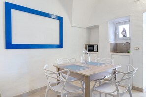 Ein Essbereich mit Holztisch und weißen Stühlen, einem blau gerahmten Bild an der Wand, einer Mikrowelle und einem Fenster mit einer aufgehängten Tasche.