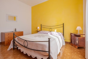 Un dormitorio con cama, cómoda de madera, mesita de noche, lámpara y espejo en la pared.
