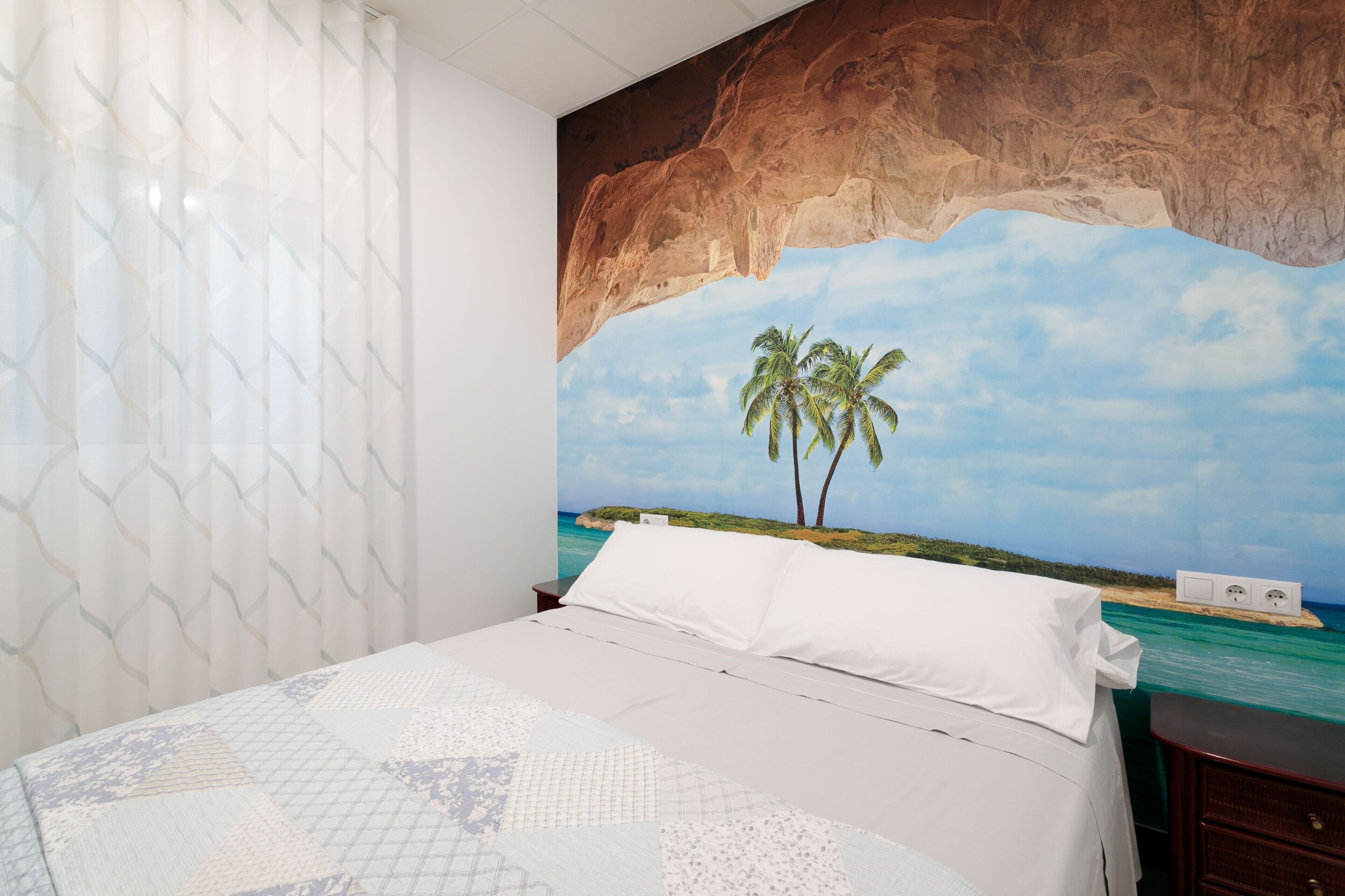 Une chambre à coucher avec un lit, une table de chevet, une fresque murale représentant une scène de plage et une fenêtre avec des rideaux.