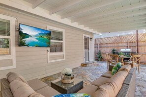 A covered patio with stone flooring, beige sofas, a flat-screen TV, and a wooden table with chairs. - Oaks Retro Cottage, Jacuzzi, Bistro Patio, Walk to Everything! (Thousand Oaks)
