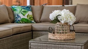 Et flettet sofabord med en flettet kurv fyldt med hvide blomster, placeret på et glastop i forbindelse med en beige sofa med et tropisk blad-mønstret pude.