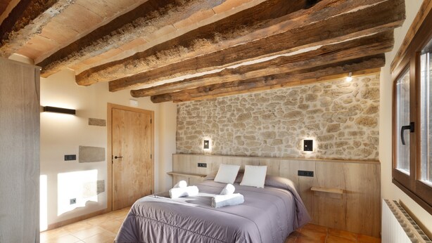 A bedroom with a stone wall, wooden beams, a bed with a purple bedspread, two bedside tables, and a window. - Rural Home 'Cal Trumfet' with Mountain View, Private Terrace and Wi-Fi (Casserres)