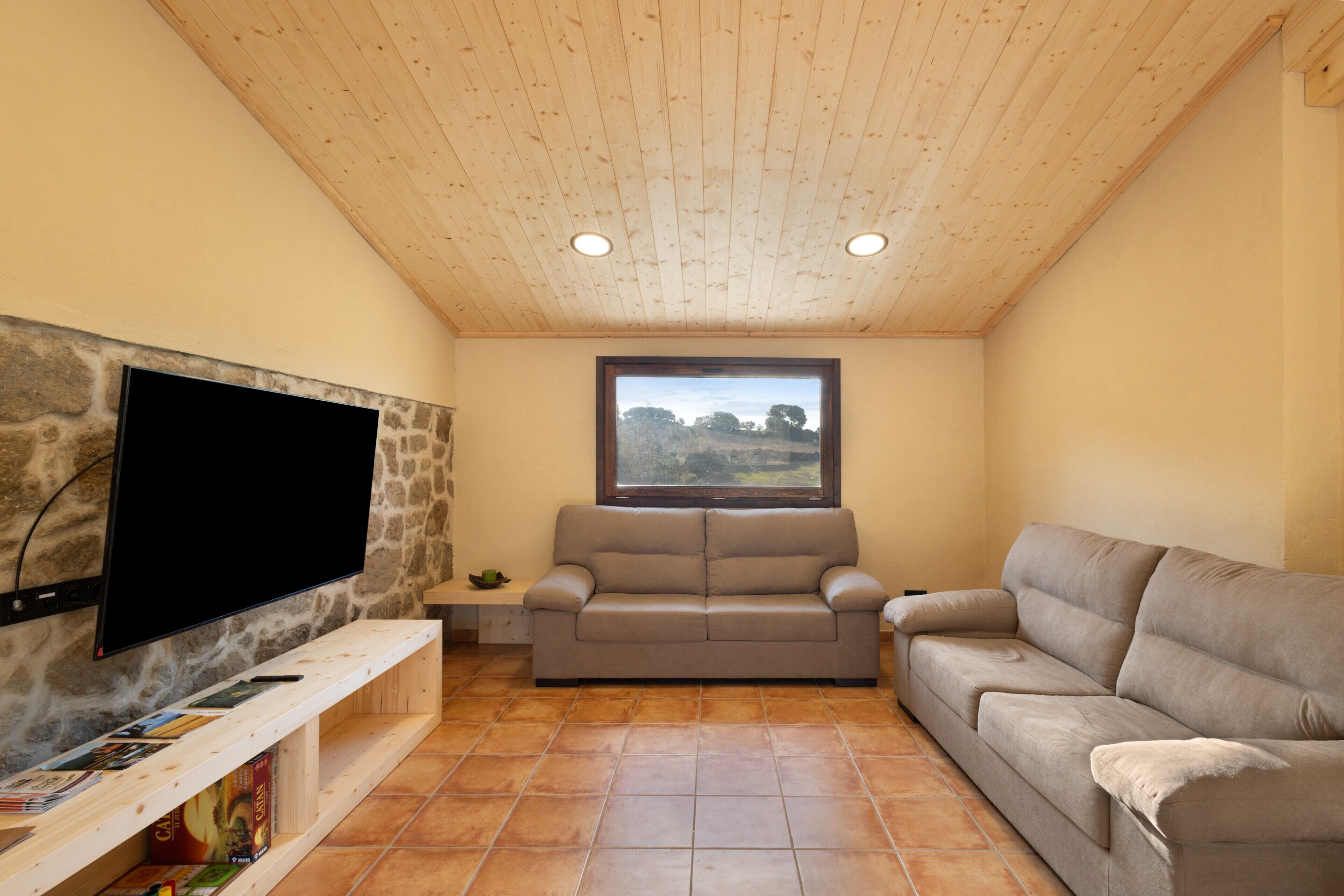 A living room with a stone wall, a flat-screen TV, a beige sofa, and a window with a view.