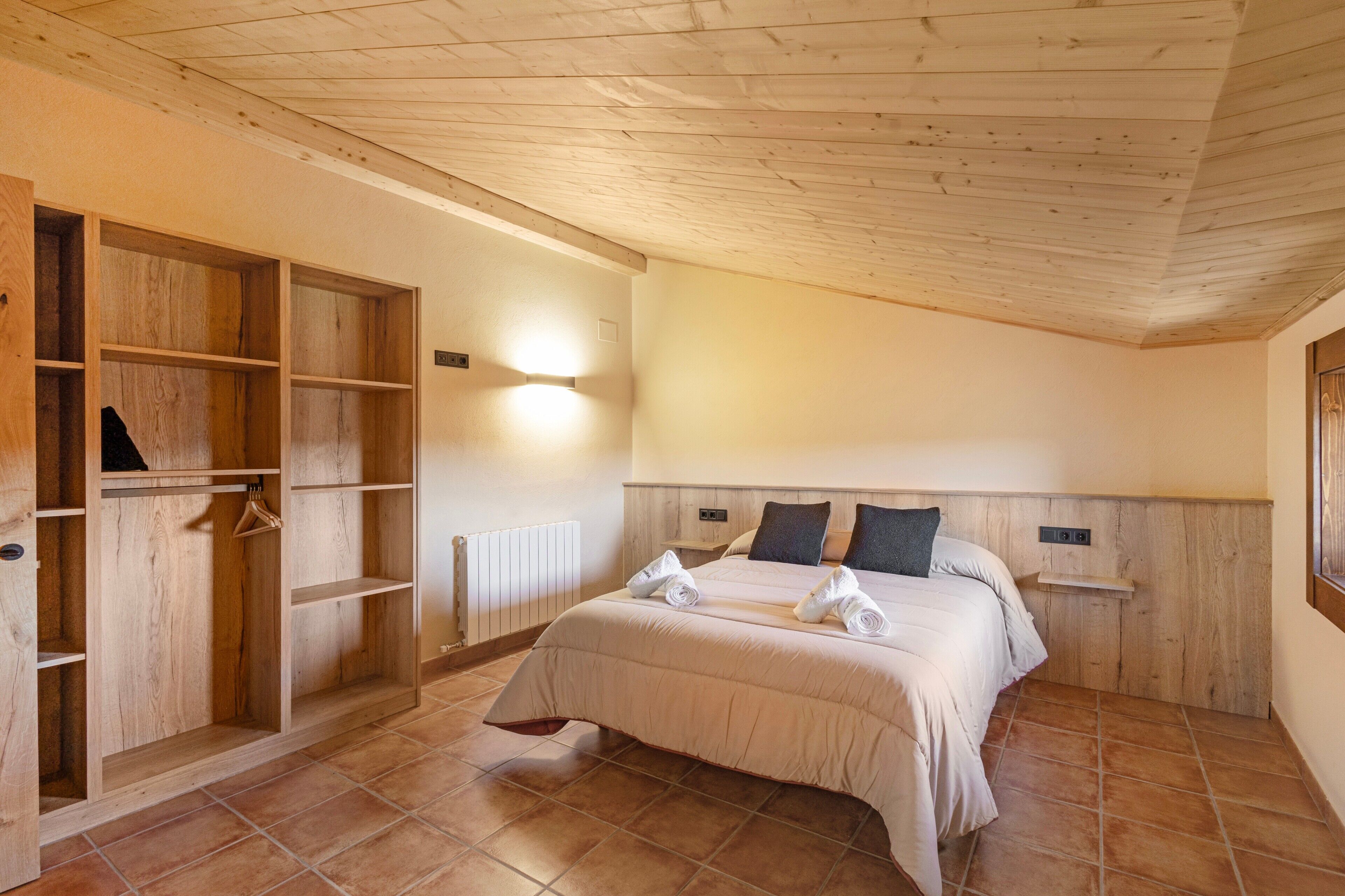 A bedroom with a wooden wardrobe, a bed with pillows, a nightstand, and a wall-mounted light.