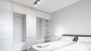Une chambre moderne avec un grand lit, une literie blanche, une table de chevet avec une lampe et un dressing avec des étagères.