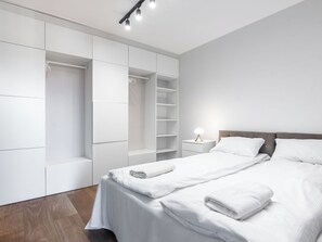 Une chambre moderne avec un grand lit, une literie blanche, une table de chevet avec une lampe et un dressing avec des étagères.