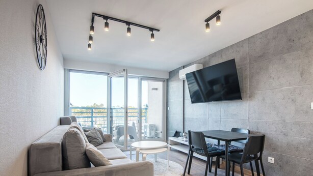 A modern living room with a sofa, dining table, chairs, a TV mounted on the wall, and a large window. - Mistral Porta Mare by Interhome (Dziwnówek)