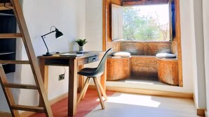 Una habitación con techo inclinado, un escritorio con silla, una escalera, una ventana con vistas y un taburete pequeño.