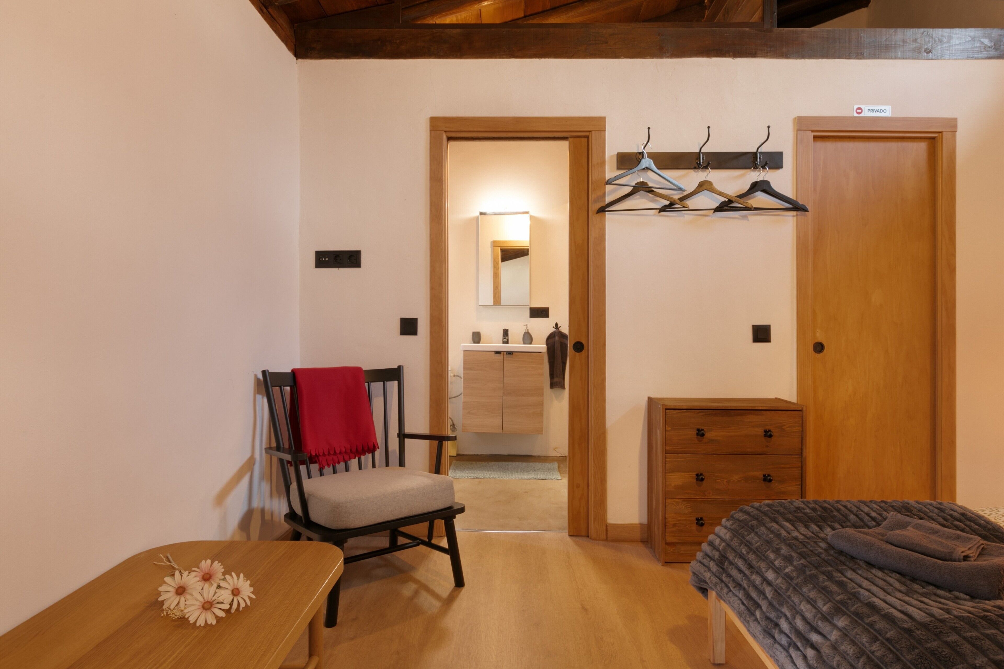 Una habitación con una cama, una silla con una manta roja, un aparador de madera y un perchero.