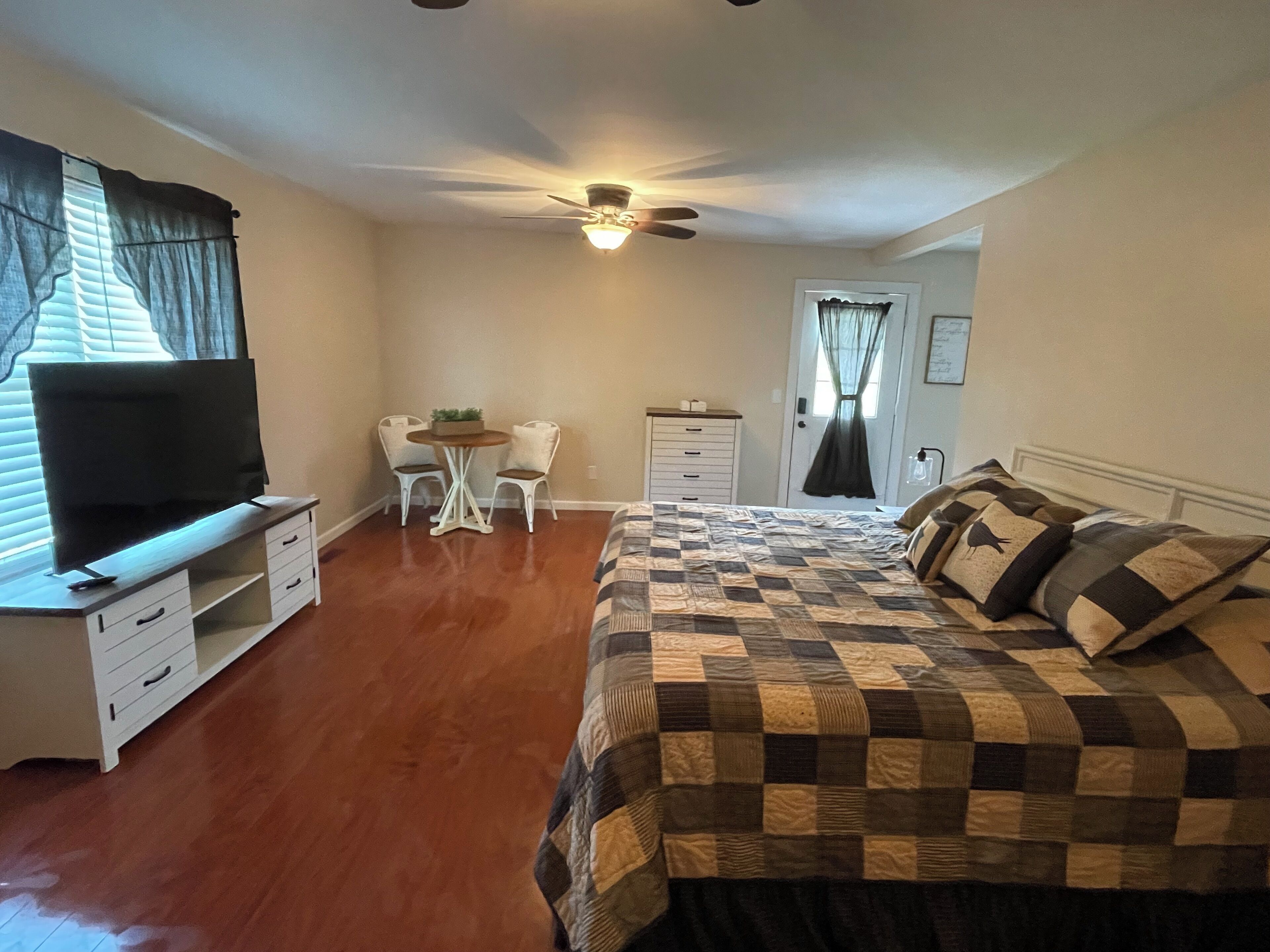 A bedroom with a bed, a TV on a stand, a desk with two chairs, a ceiling fan, and a door with curtains.