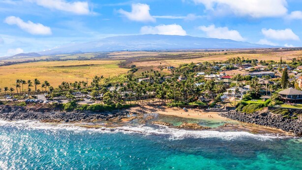 Un complejo turístico frente al mar con propiedades en la playa, palmeras y vistas a montañas distantes.