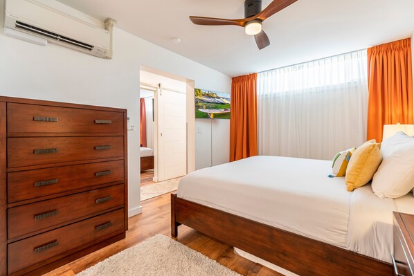 A bedroom with a wooden bed, a ceiling fan, an air conditioner, and a view of a balcony through a window with orange curtains.