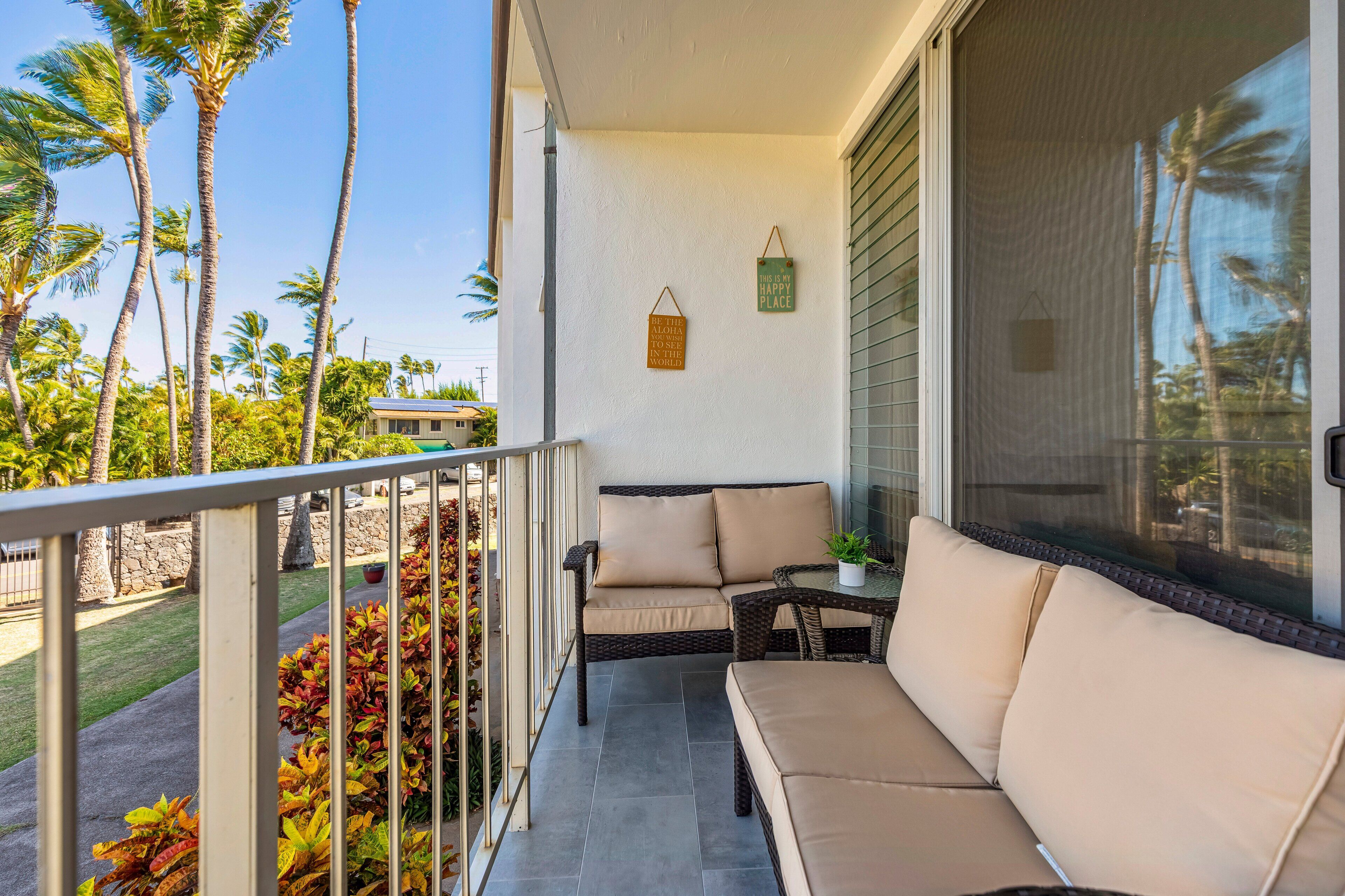 Un balcon avec un canapé et une table, des palmiers et une maison en arrière-plan.
