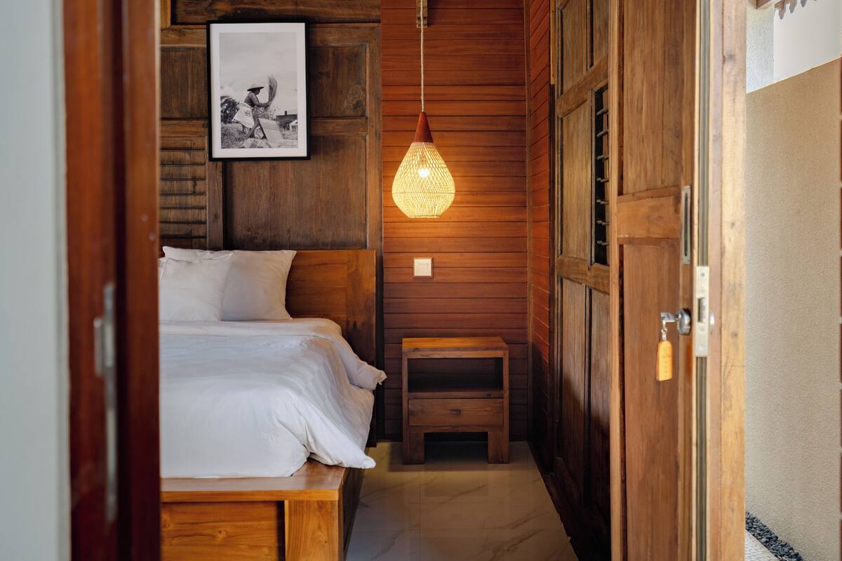 A wooden room with a bed, a small wooden bench, a hanging light fixture, and a framed picture on the wall.
